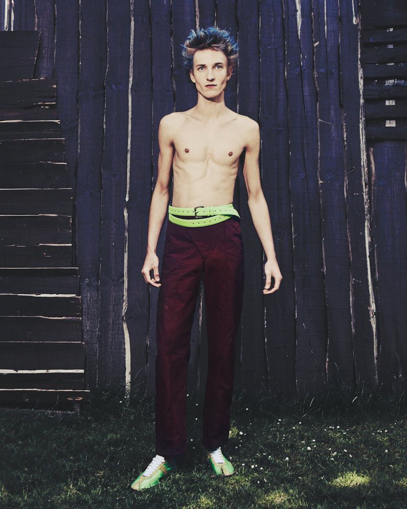 Full-body portrait of a slender shirtless adult male with dyed blue hair, wearing red pants and a green belt, standing outdoors.