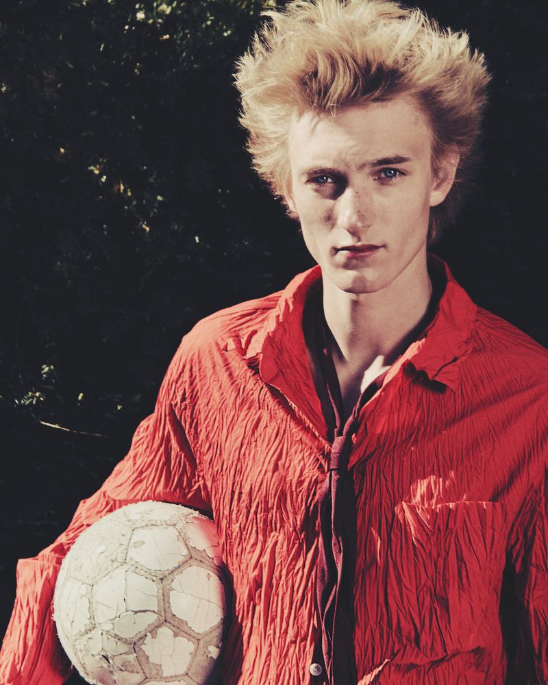 Young fair-skinned male with blond hair, wearing an orange shirt and tie, holds a cracked soccer ball in an outdoor setting against a dark background.