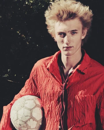 Young fair-skinned male with blond hair, wearing an orange shirt and tie, holds a cracked soccer ball in an outdoor setting against a dark background.