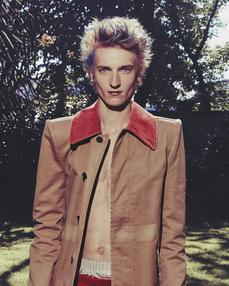 A person with dyed hair and a tan jacket stands in a grassy area with trees, looking directly at the camera in a medium shot.
