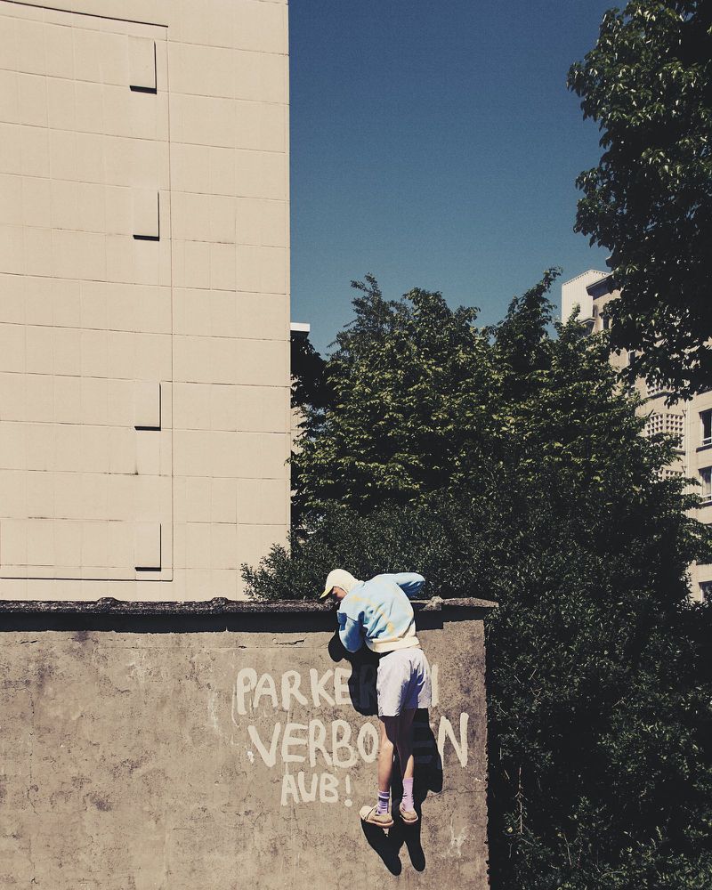 Man climbs a wall with graffiti in an urban environment on a sunny day, with trees and buildings in the background against a clear blue sky.