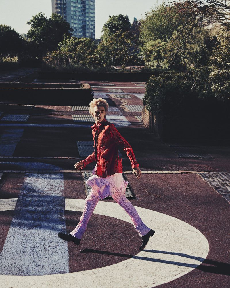Fashionable young adult in a red shirt and pink pants strides purposefully across a concrete urban landscape on a sunny day.