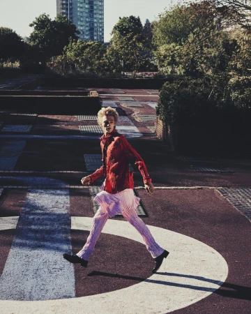 Fashionable young adult in a red shirt and pink pants strides purposefully across a concrete urban landscape on a sunny day.