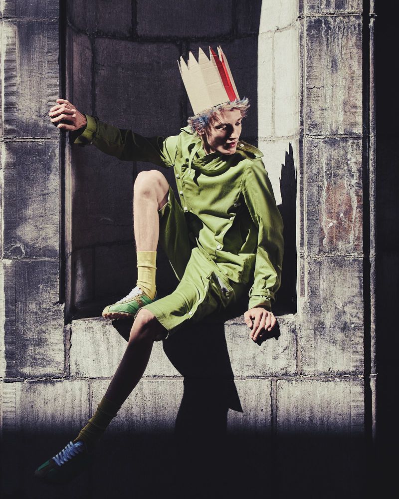 Young adult in a green suit and cardboard crown sits in a stone archway bathed in light and shadow with an expressive, cool fashion aesthetic.