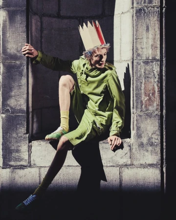 Young adult in a green suit and cardboard crown sits in a stone archway bathed in light and shadow with an expressive, cool fashion aesthetic.