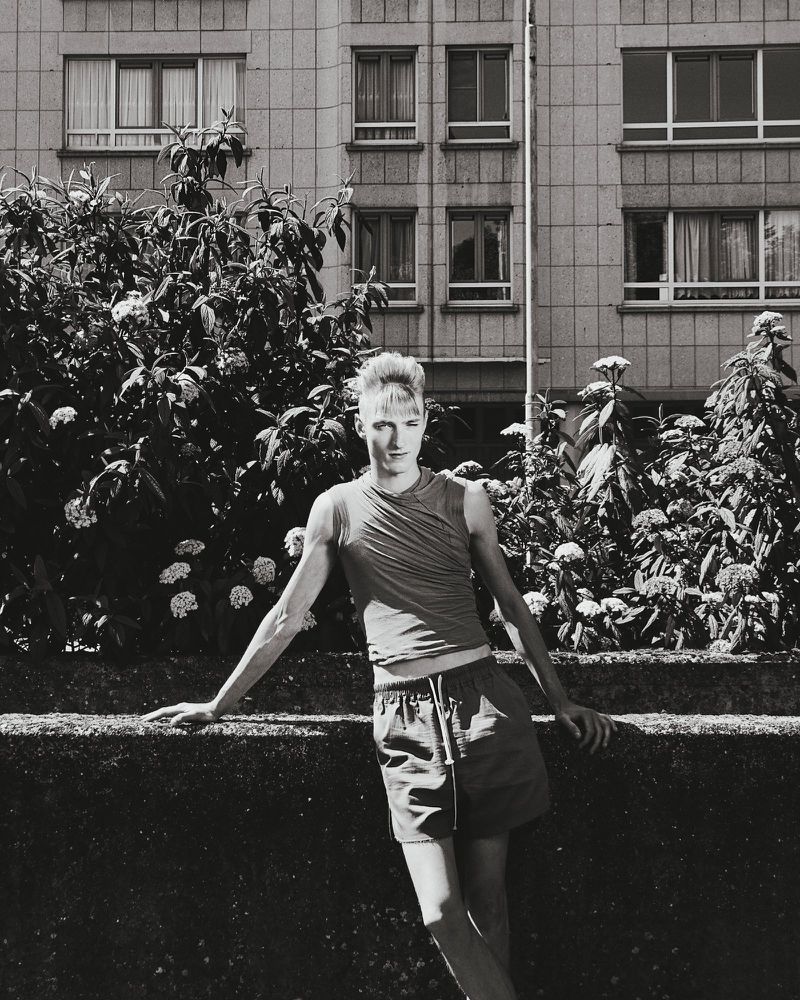 Monochrome shot of a young adult leaning against a wall with lush green plants, a building with windows in the background.
