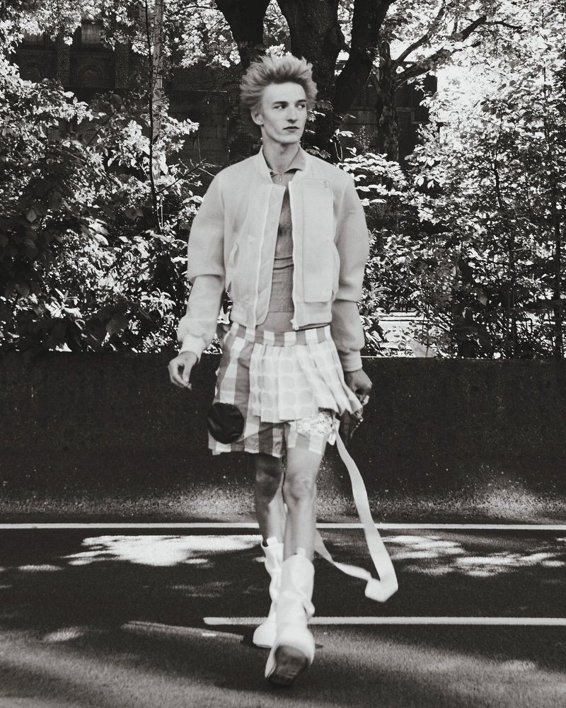 Young adult with spiky hair and jacket walks on road wearing a plaid skirt and white boots; trees are in the background, black and white.