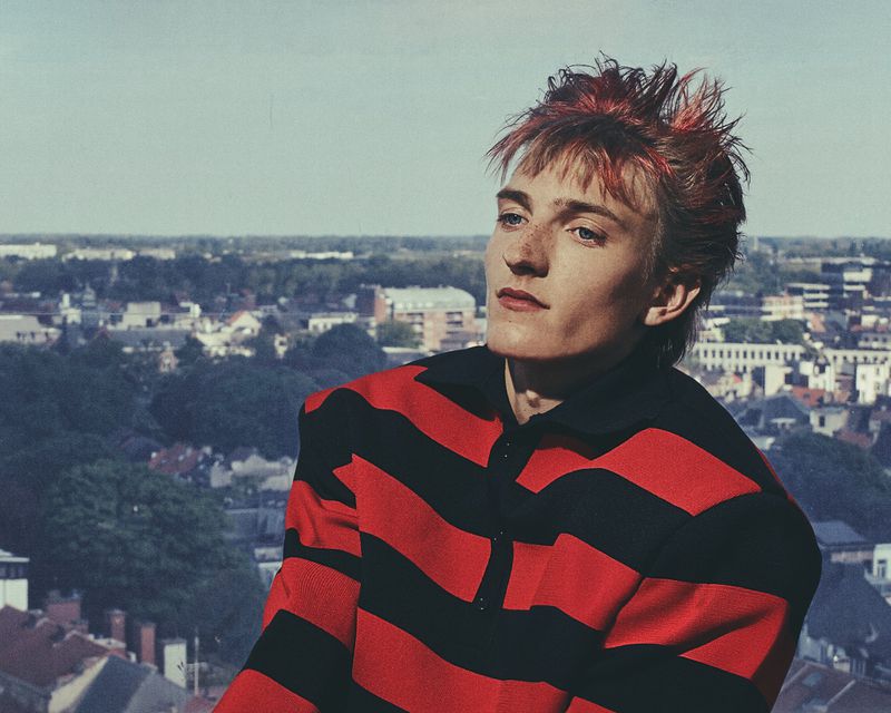 Portrait of a young adult with dyed red hair and a red and black striped shirt, gazing off camera with an urban backdrop.