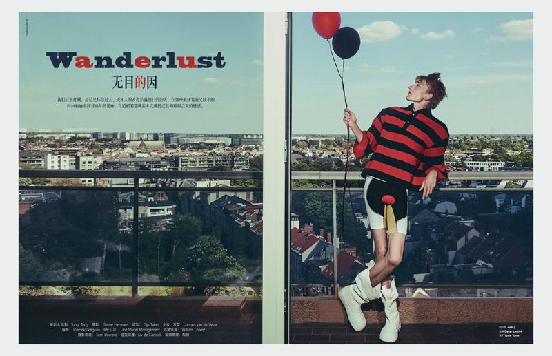 Fashion editorial shot of a young adult man in a striped shirt and shorts, holding balloons on a rooftop overlooking a city.