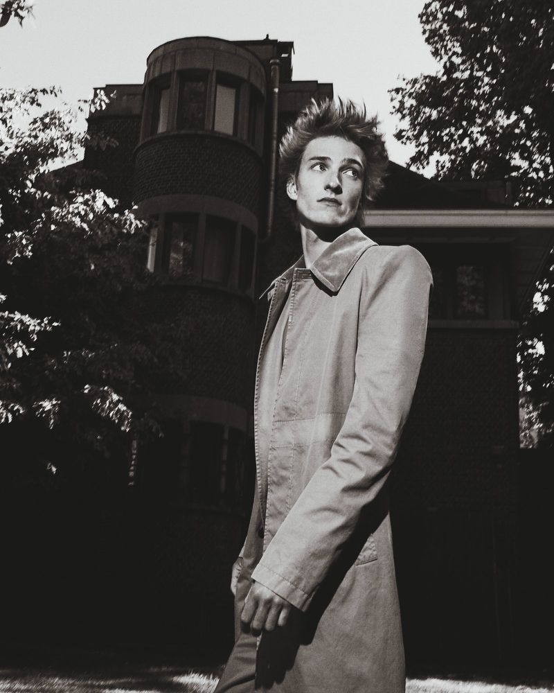 Monochrome portrait of a fair-skinned adult man in a light coat, with spiky hair, outside a brick building in a garden setting.