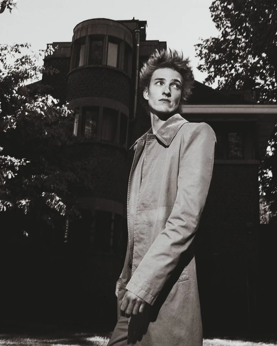 Monochrome portrait of a male model wearing a long coat standing outdoors in front of a residential building.