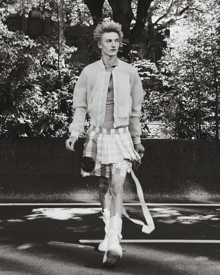 Model walking outdoors on a road wearing a bomber jacket and patterned shorts in a black and white editorial image