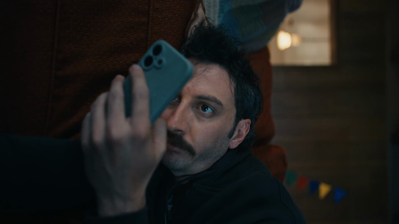 Man with a mustache holds a phone up to his face indoors, with blurred background details and soft lighting in a relaxed environment.