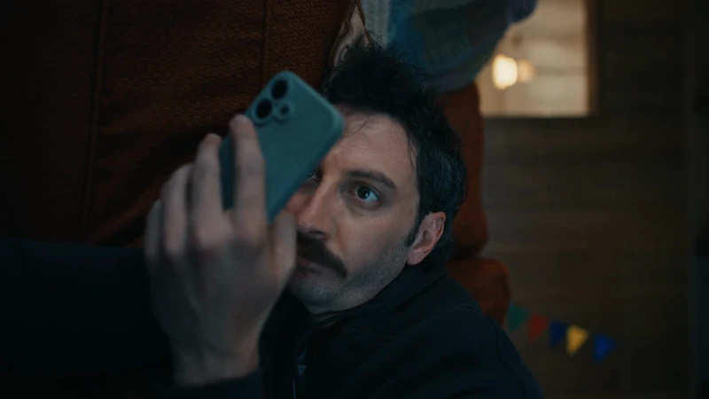 Man with a mustache holds a phone up to his face indoors, with blurred background details and soft lighting in a relaxed environment.