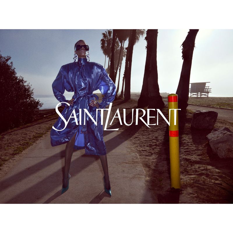 Fashion editorial shot of a woman in a blue coat posing on a beach path with palm trees and a lifeguard station in the background.