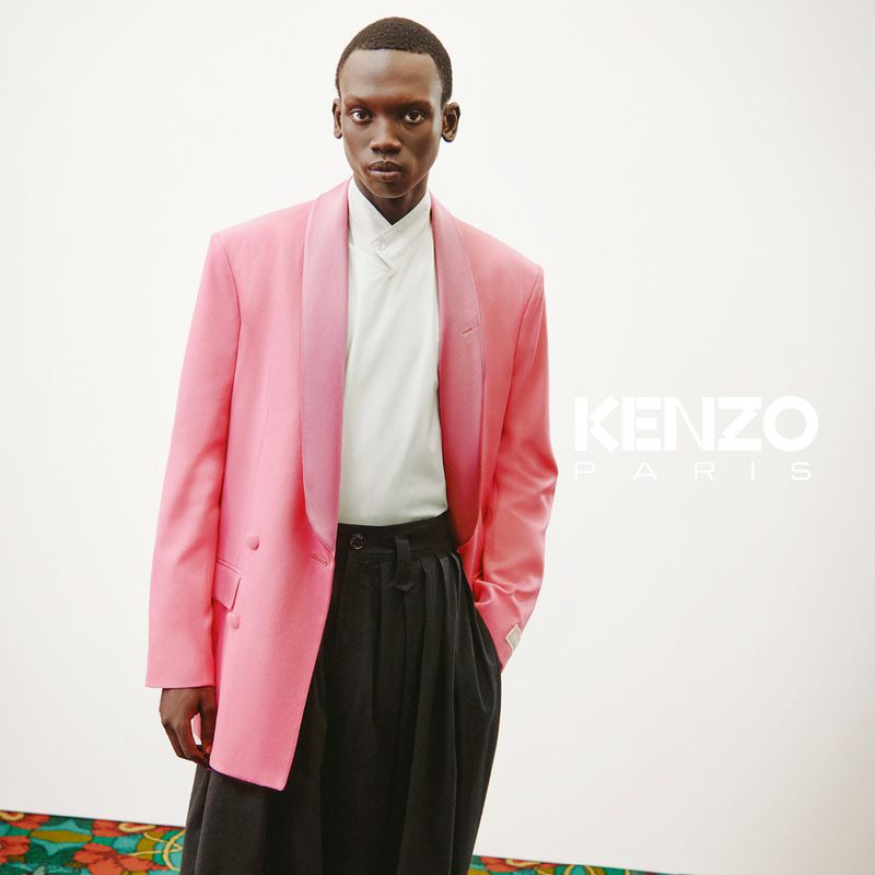 A slender African-American male model poses indoors wearing a pink blazer, white shirt, and black pants by Kenzo Paris.