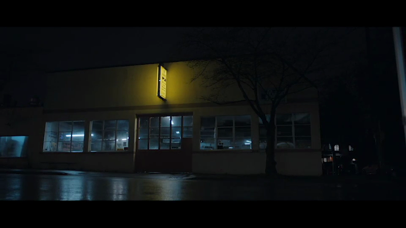 Exterior of a building at night with lit windows and a tree. The scene is dark with wet pavement reflecting lights.