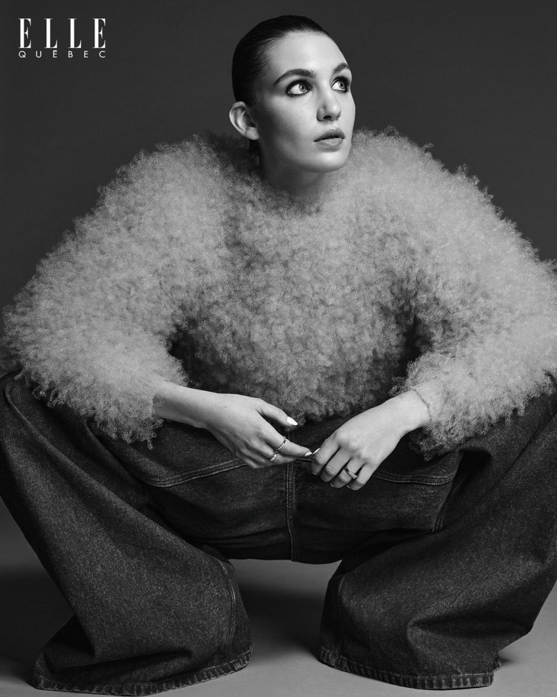 Monochrome portrait of a woman in oversized denim and a textured top, squatting in a studio setting with ELLE Quebec logo visible.