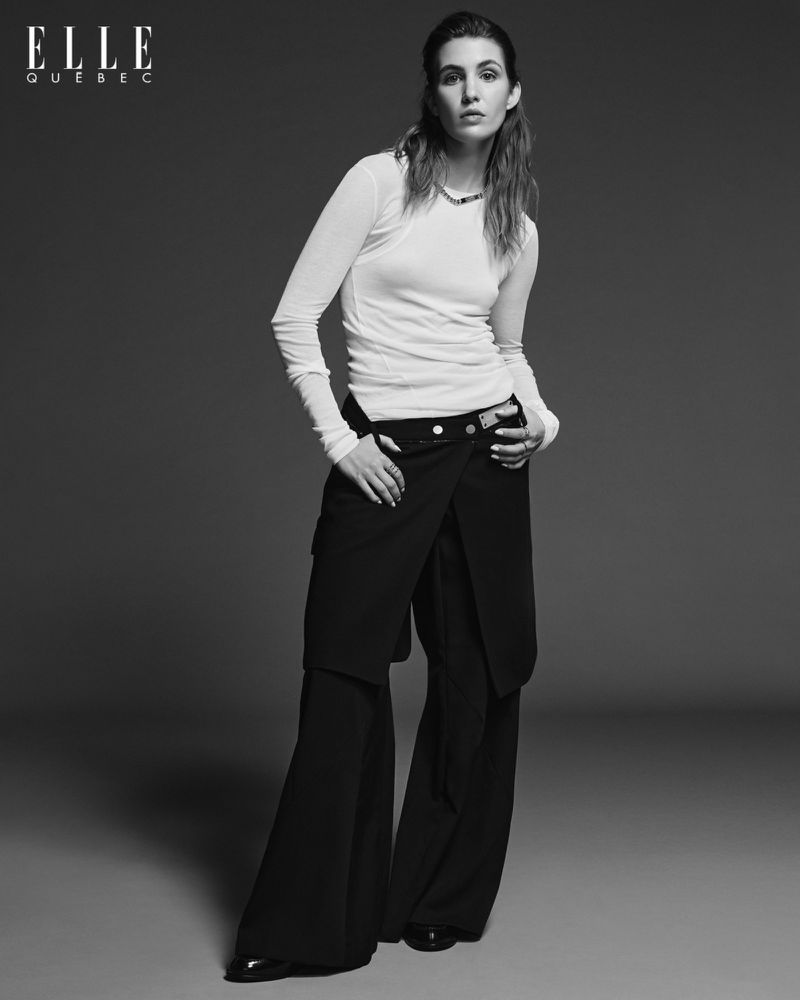 A person in a white long-sleeve shirt and black flared pants poses in front of a grey backdrop with the Elle Quebec logo visible.