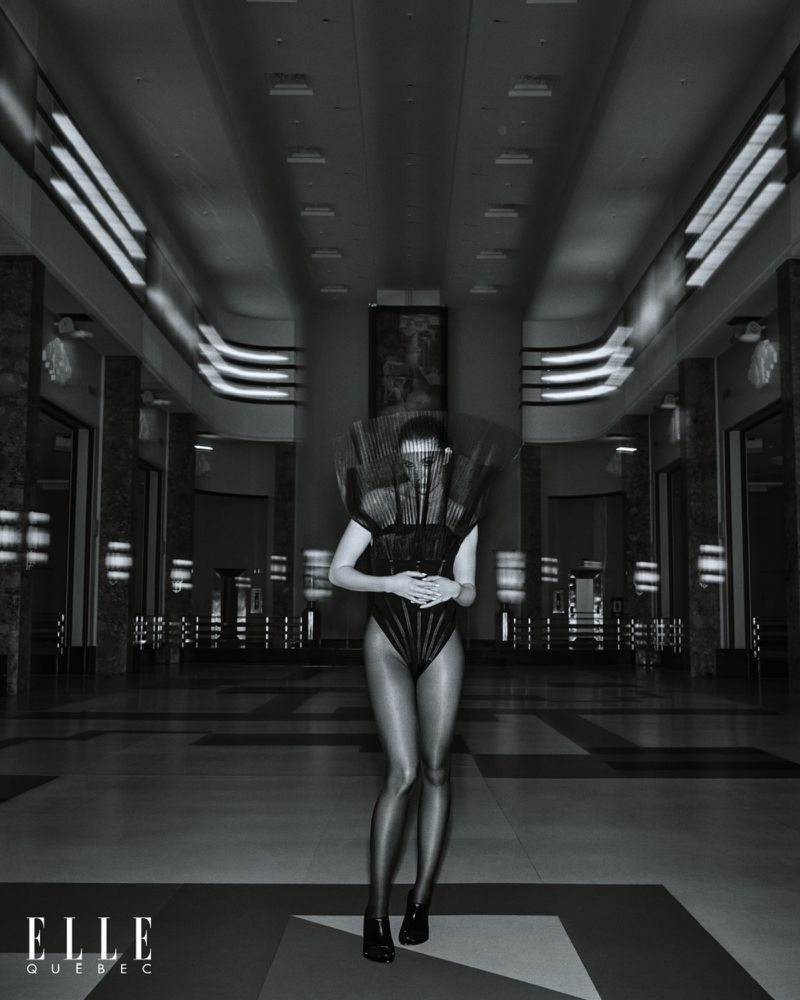 Monochrome full-shot of a woman wearing a futuristic bodysuit and headdress, posing in an indoor hall with architectural elements.