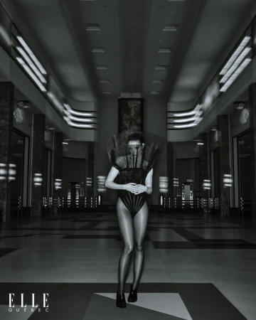 Monochrome full-shot of a woman wearing a futuristic bodysuit and headdress, posing in an indoor hall with architectural elements.