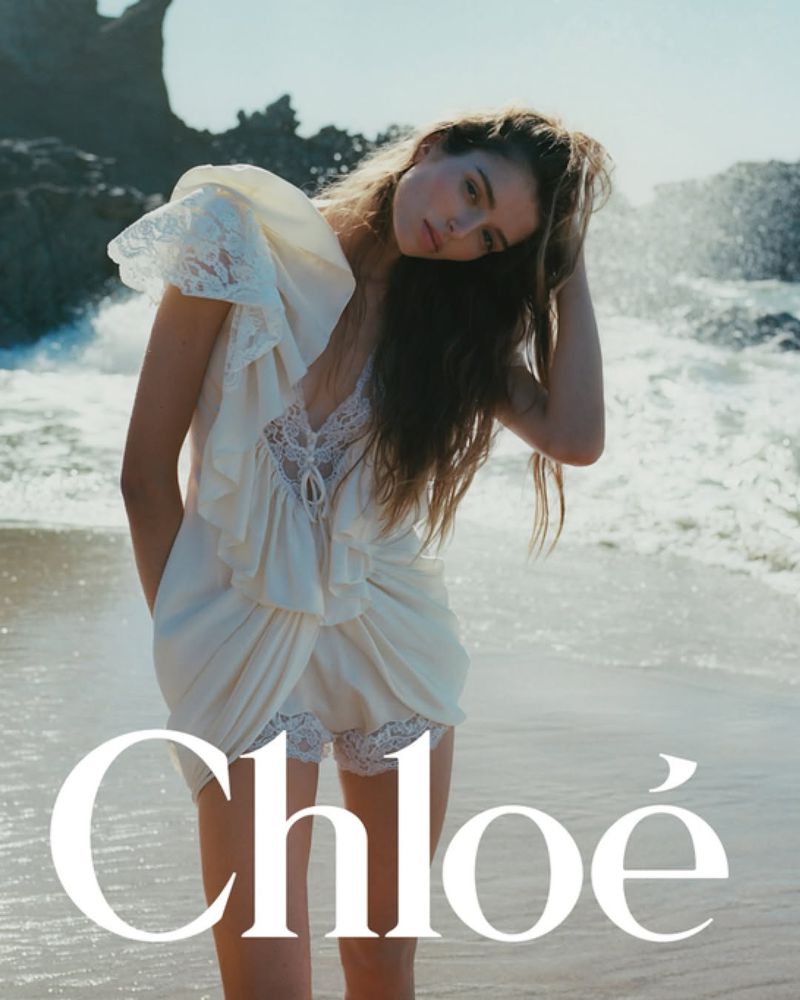 A woman in a white dress poses on a beach with ocean waves and rocks in the background, bathed in soft daylight, fashion editorial.