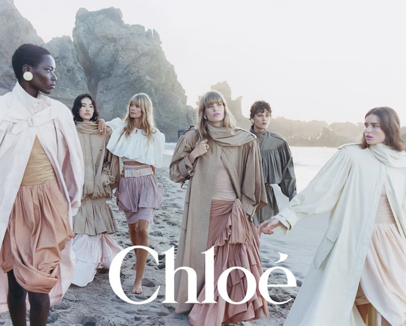 Fashion editorial featuring a group of women in flowing dresses and coats posing on a beach with rock formations and soft, natural light.