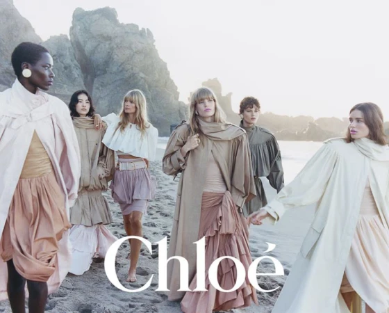 Fashion editorial featuring a group of women in flowing dresses and coats posing on a beach with rock formations and soft, natural light.