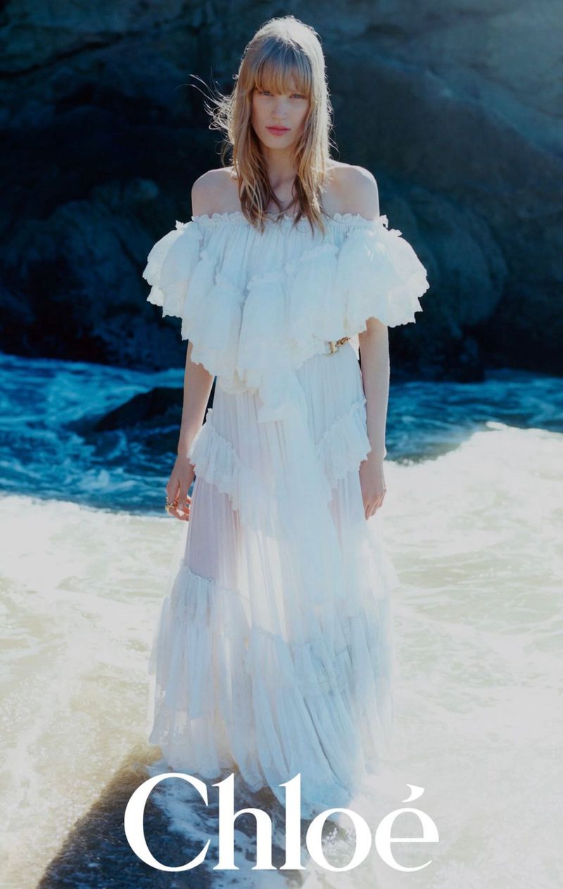 A fashion shot shows a blonde adult female model posing in a long white dress at the beach, with rocks and ocean water in the background.