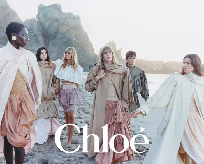 Group of six female models wearing layered neutral and pastel garments standing on a sandy beach.