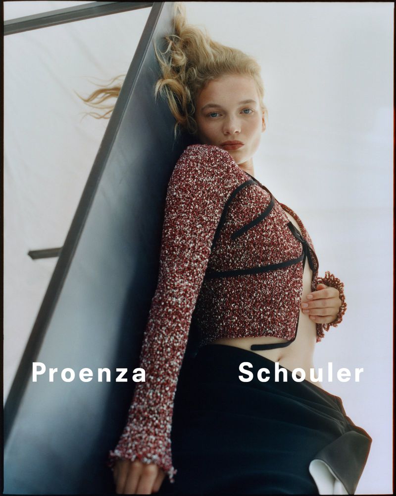 Fashion shot of a woman with blond hair wearing a patterned burgundy sweater with black pants against a wall, with brand text overlay.