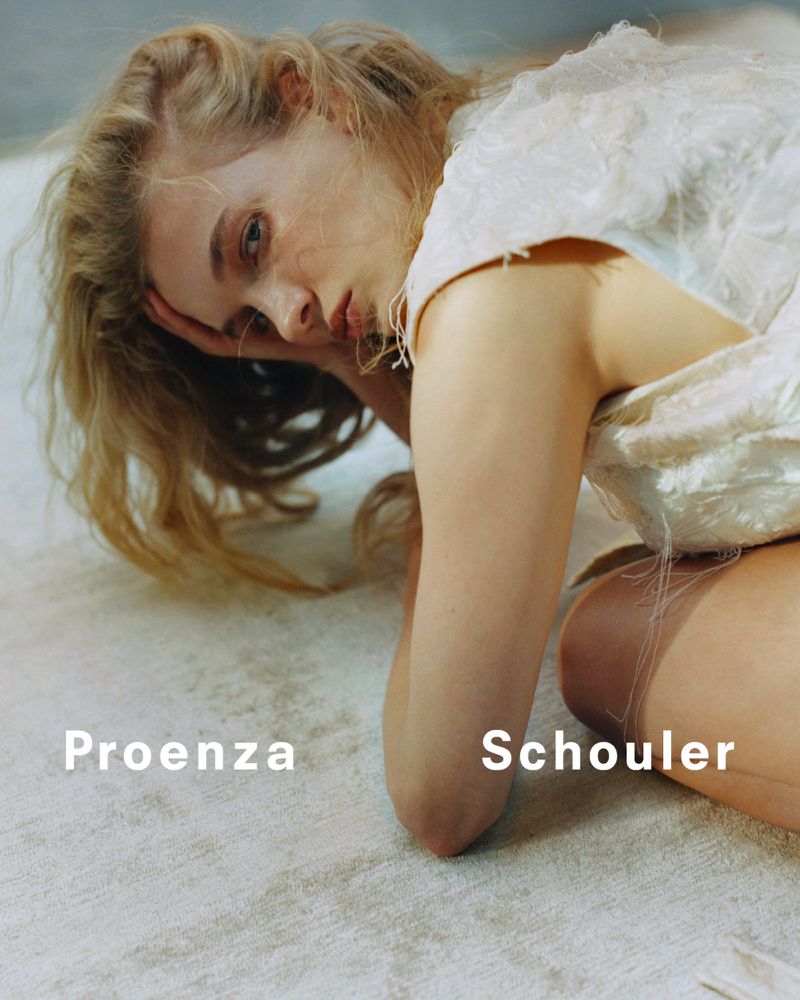 Fashion shot of a blonde woman in a white dress lying on a textured white floor. She rests her head in her hand and looks to the side.