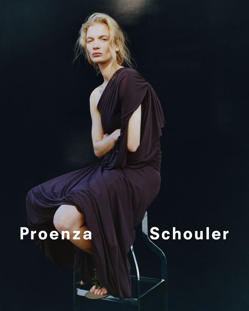 A model poses while sitting on a stool in a Proenza Schouler dress against a dark background in a fashion studio shot.