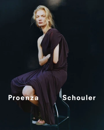 A model poses while sitting on a stool in a Proenza Schouler dress against a dark background in a fashion studio shot.