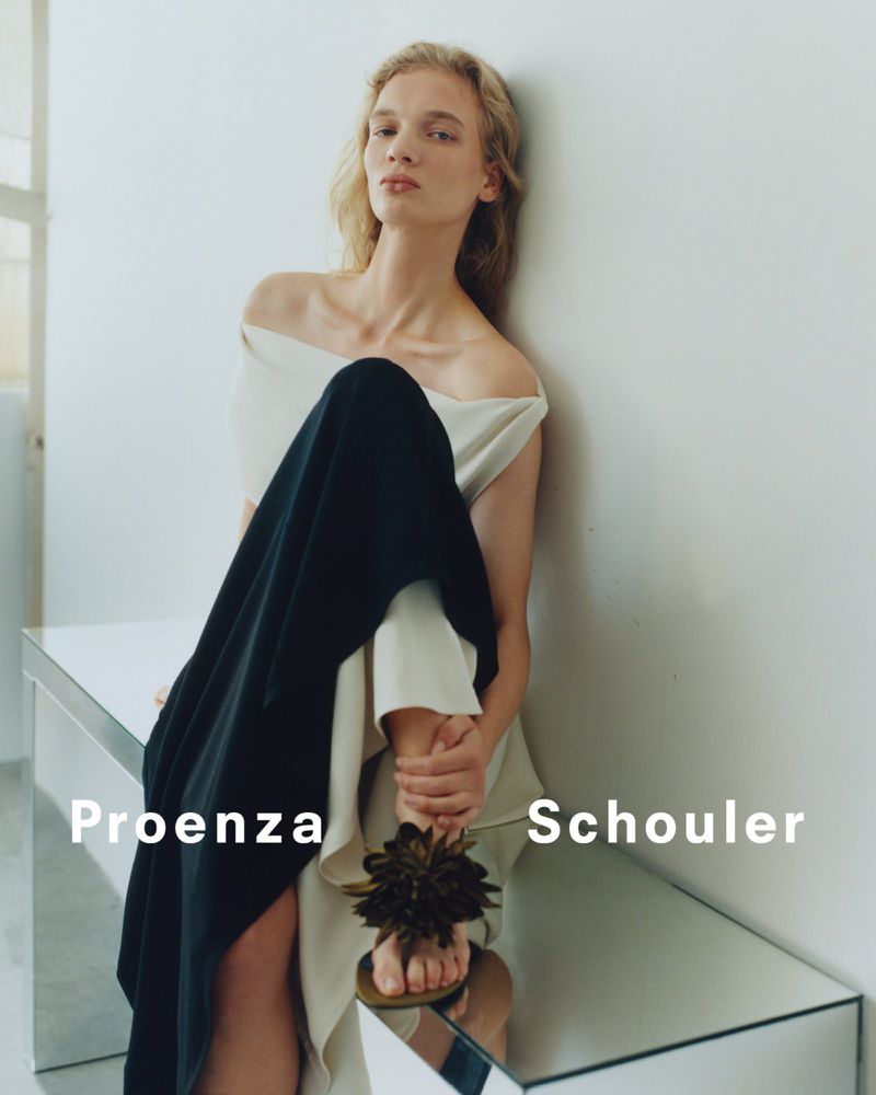 Fashion editorial shot of a woman wearing an off-the-shoulder top and black skirt sitting on a reflective table indoors.