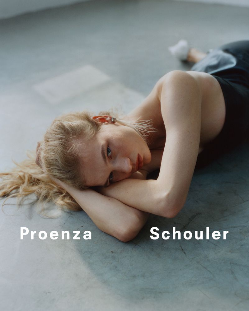 Fashion shot of a blonde woman lying on a gray surface with her arms crossed under her head, looking at the camera with neutral expression.