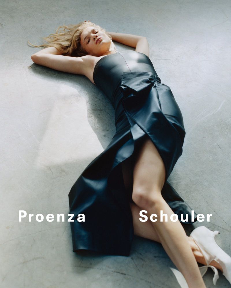 Fashion shot of a blonde woman lying on her back wearing a black leather dress and white shoes in a studio with grey flooring.