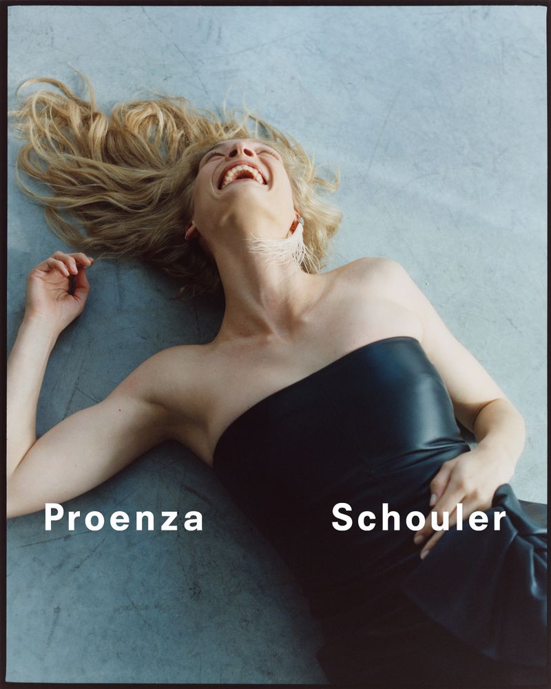 An adult woman with blonde hair smiles while lying on a smooth gray surface wearing a strapless black dress in a studio shot.