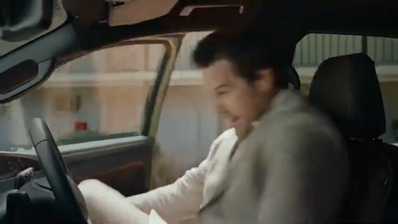 Adult male in light clothing sits inside a car, looking down with a smiling expression. Steering wheel is visible in the car interior.