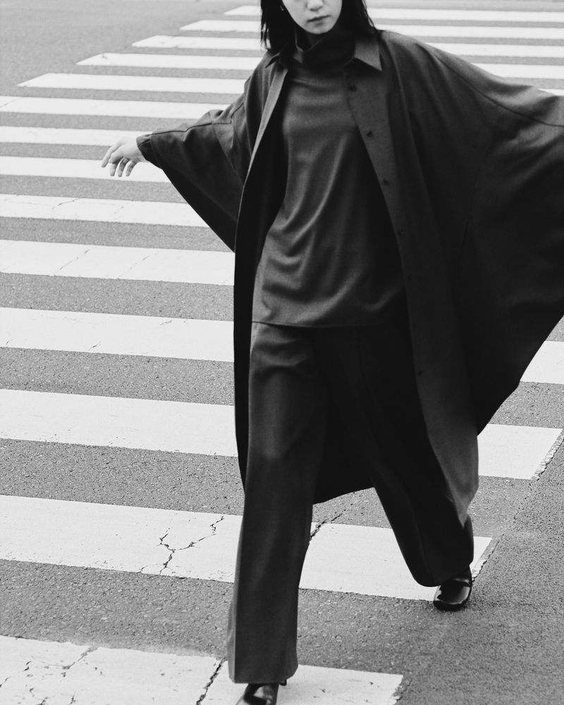 Monochrome fashion editorial image of a woman crossing a pedestrian crosswalk wearing an oversized coat and trousers.