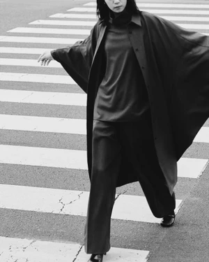 Monochrome fashion editorial image of a woman crossing a pedestrian crosswalk wearing an oversized coat and trousers.