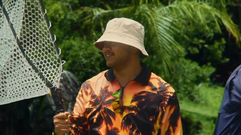 Adult man wearing a bucket hat and tropical print shirt holds a fishing net in a lush, green outdoor environment with palm trees.