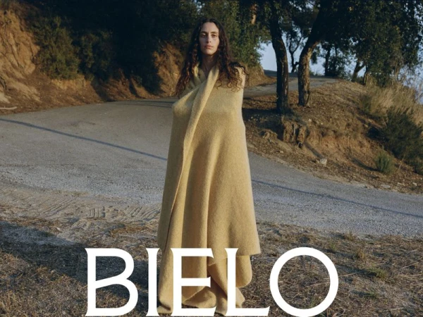 A young adult woman stands outside wrapped in a yellow blanket, set against a landscape with trees and a winding asphalt road.