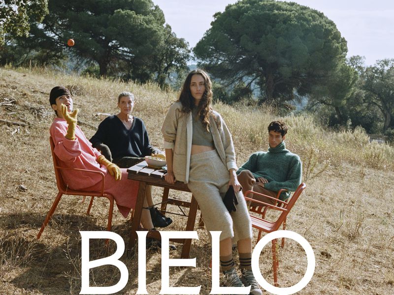 Outdoor fashion shot features four people seated around a table in a field of dry grass, trees in background, 'BIELO' text overlay.