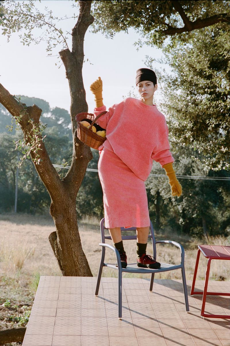 A woman in a pink outfit poses on a chair outdoors with a basket of lemons, trees in the background, and natural lighting.