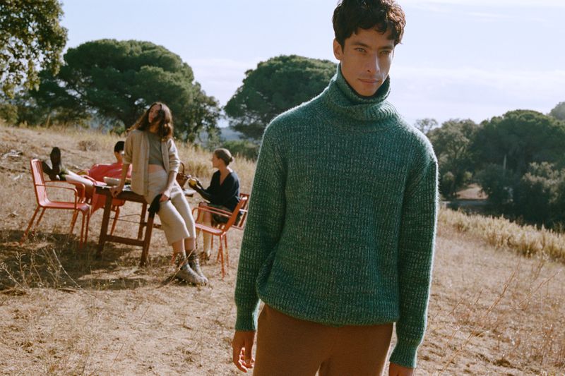 Portrait of a young man wearing a green turtleneck sweater standing outdoors, with people seated at tables in the background.