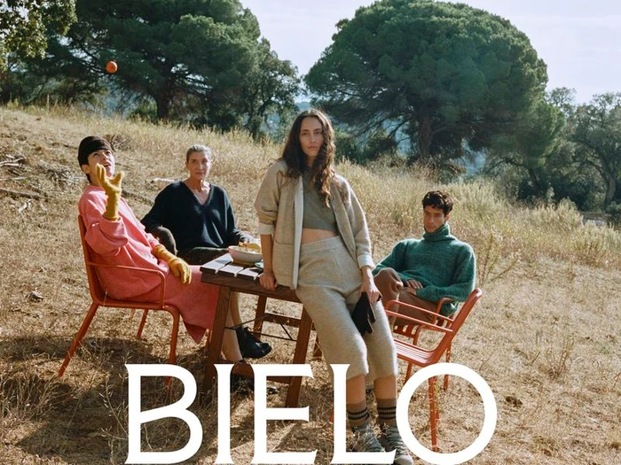 Four models in knitwear gathered around an outdoor wooden table in a grassy landscape with pine trees for Bielo AW26.