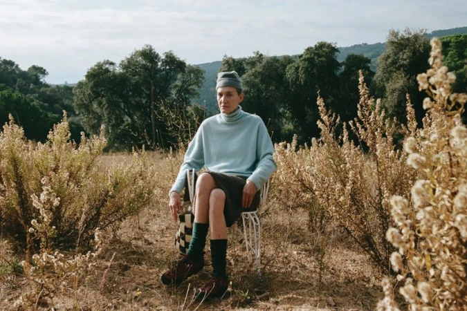 An older woman in a pale blue turtleneck and grey beanie seated on a white wire chair in a dry grassy meadow.