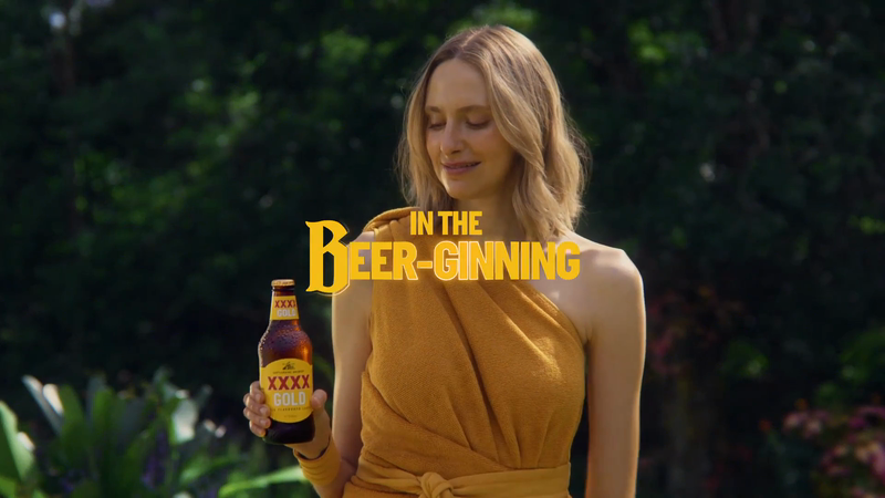 Adult woman holds a bottle of XXXX Gold beer in a lush green outdoor setting; text overlay 'In The Beer-ginning.'