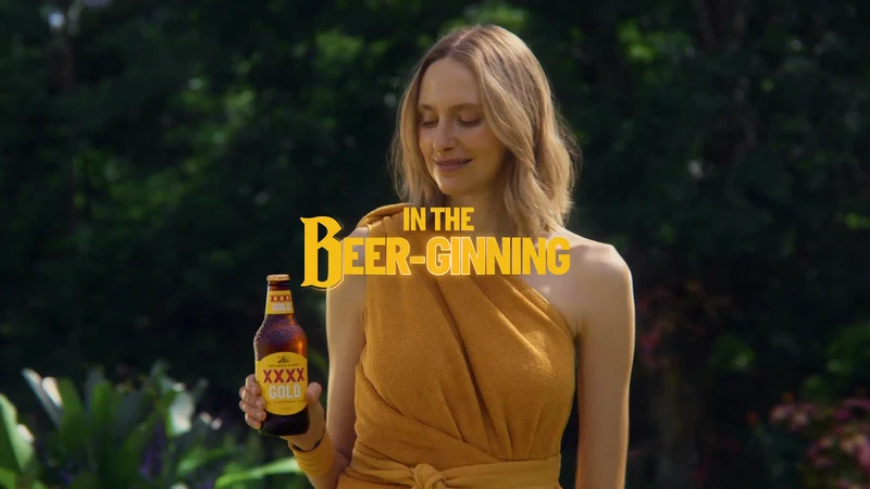 Adult woman holds a bottle of XXXX Gold beer in a lush green outdoor setting; text overlay 'In The Beer-ginning.'